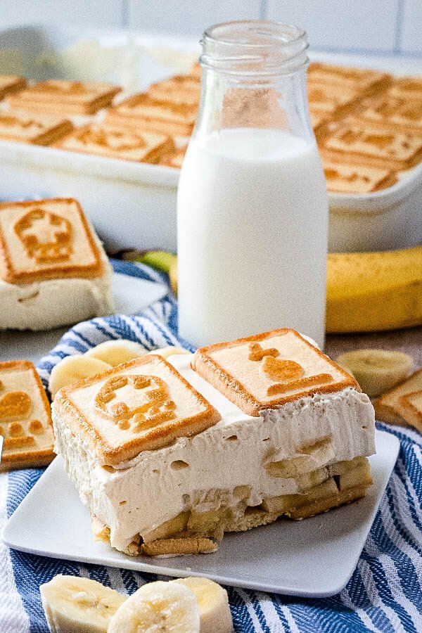 Budín de plátano en un plato con un vaso de leche y una bandeja de postre en segundo plano.