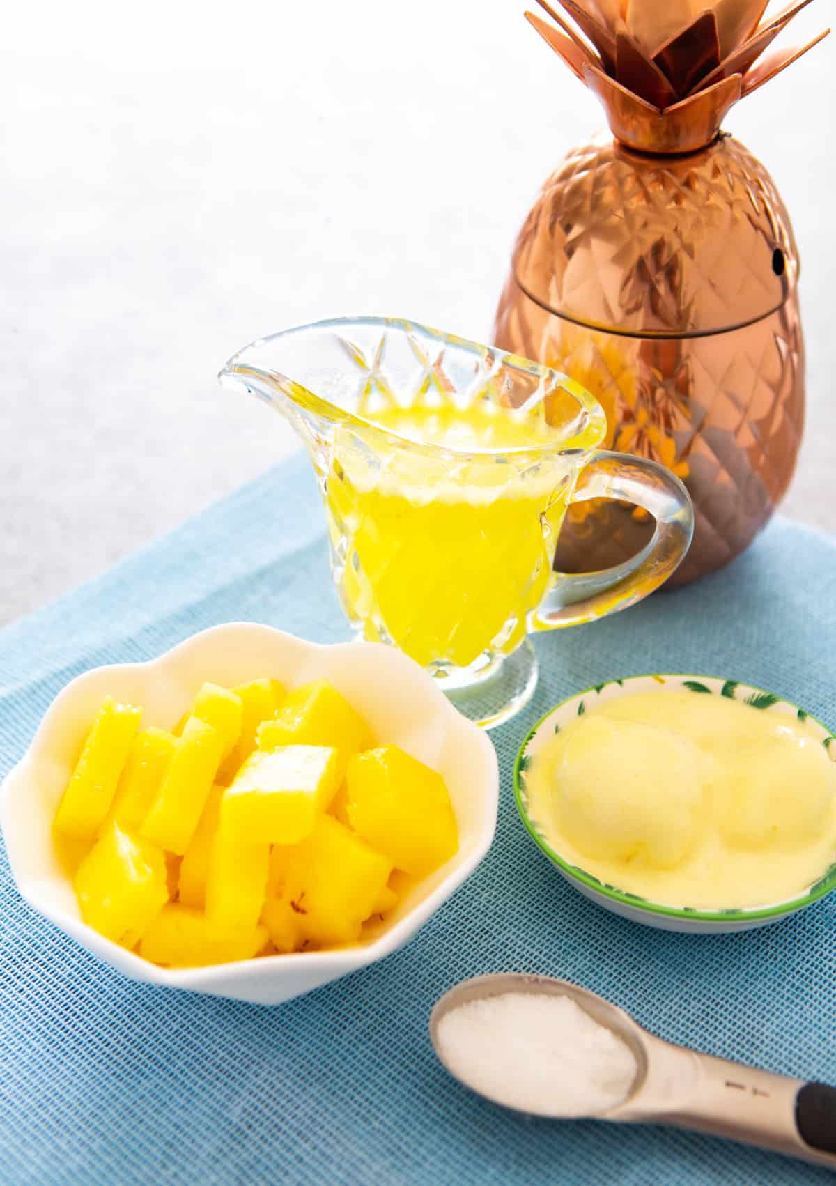 Piña congelada en un tazón, jugo en una jarra y helado de vainilla, todo sobre una mesa.