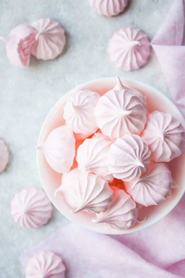 Imagen de arriba de galletas de merengue rosa bebé en un tazón blanco con tela rosa.