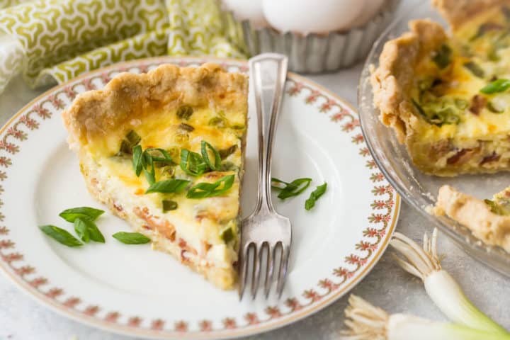 Rebanada de quiche Lorraine en plato de porcelana con tenedor de plata y chalota fresca en rodajas.