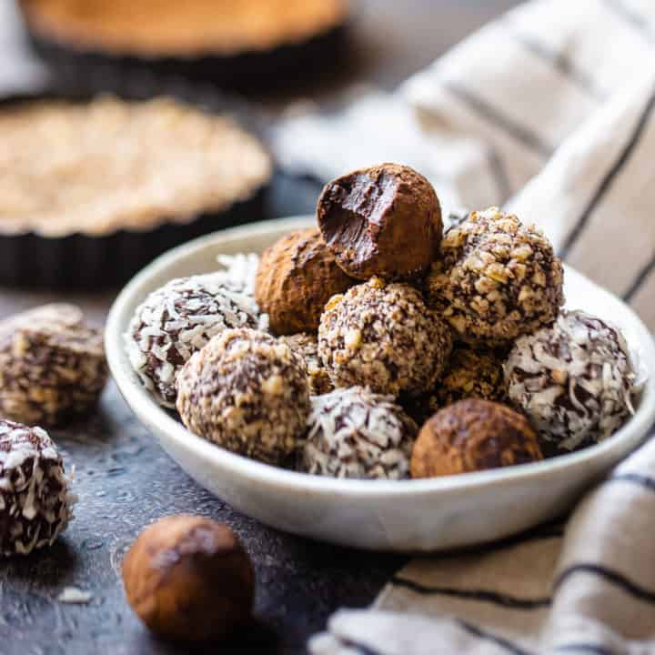 Trufas de chocolate apiladas en un plato de cerámica con cajas de nueces picadas al fondo.