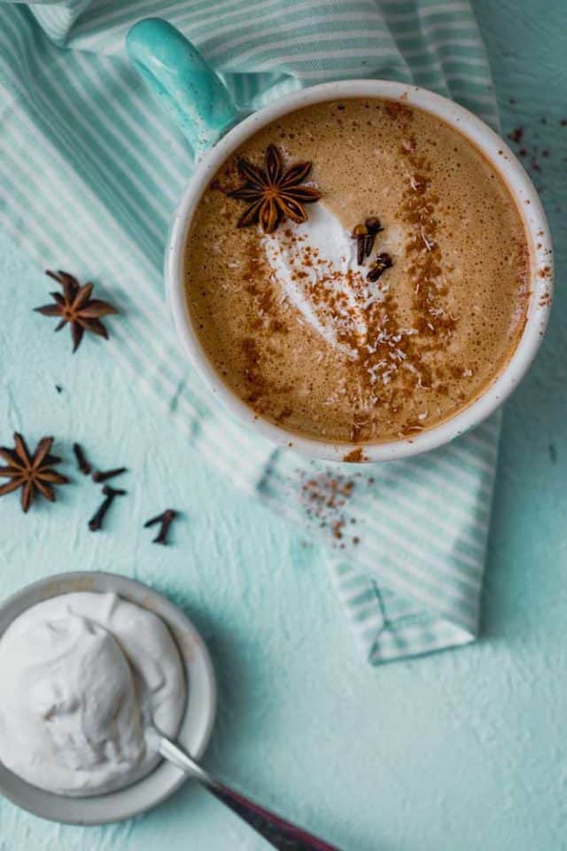 Primer plano de café mezclado con aceite de coco, que muestra una taza espumosa y cremosa con una servilleta azul debajo. También hay anís estrellado y clavo de olor entero como guarnición, así como crema de coco y una pizca de canela en polvo.