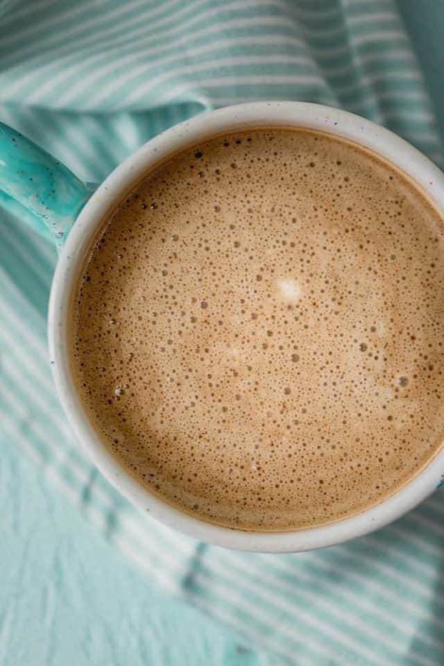 Primer plano de café mezclado con aceite de coco, que muestra una taza espumosa y cremosa con una servilleta azul debajo