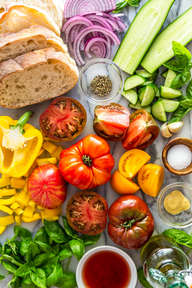 Un hermoso arco iris de ingredientes para ensalada Panzanella: pan, cebolla roja, pepino, tomate, pimiento amarillo, albahaca, vinagre de vino tinto, aceite de oliva, mostaza Dijon, sal, pimienta.