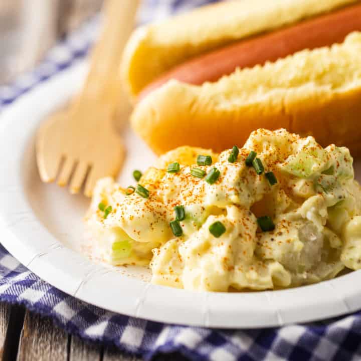 Una cucharada de ensalada de patata en un plato de papel con un perrito caliente al fondo.